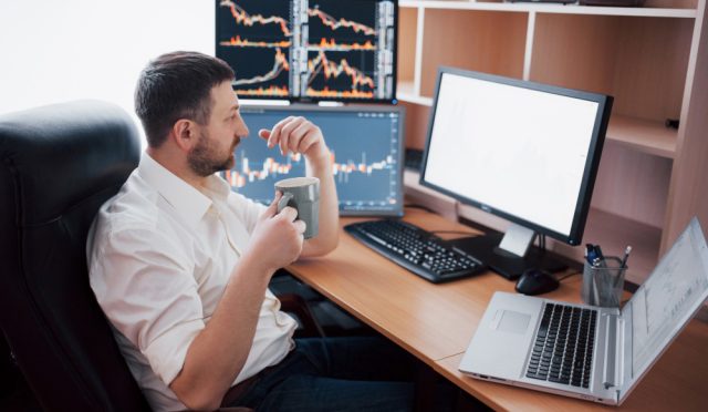 young-businessman-is-sitting-office-table-working-computer-with-many-monitors-diagrams-monitor-stock-broker-analyzes-binary-options-charts-hipster-man-drinking-coffee-studying